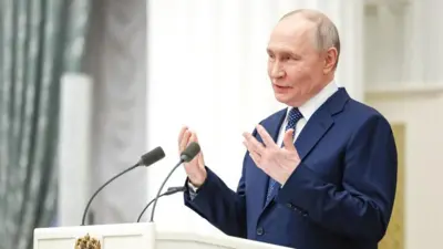 Prezydent Putin w białej koszuli, niebieskim krawacie w kropki i niebieskim garniturze, gestykuluje rękami, przemawiając na białym podium z dwoma mikrofonami. Po lewej stronie zdjęcia znajduje się jasnozielona, ​​wzorzysta zasłona.