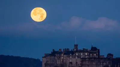 The full October moon, known as the Hunter's sets behind Edinburgh Castle, Scotland. 17 October 2024. The moon is on the left hand side of a blue dusk sky. The castle in in the foreground to the right.