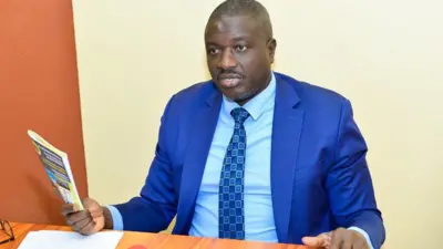 Faustin Ndikumana yambaye kositime n&#x27;ikaruvati yy&#x27;ubururu, n&#x27;ishati igomba kwera, afise igitabo mu kuboko kw&#x27;i buryo