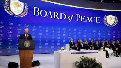 Kürsüde beyaz saçlı bir adam. İleride yan yana sandalyelerde çeşitli kadın ve erkekler. Hepsi sahnede. Arkadaki mavi fonda Barış Kurulu yazıyor.