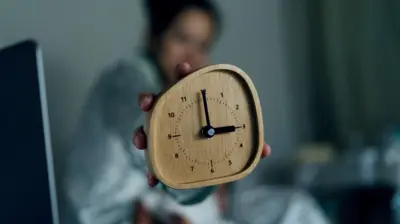 Pessoa sentada na cama mostra um despertador marcando três horas