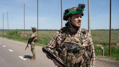 На дорозі серед відкритої місцевості стоїть військовий у тактичному спорядженні, тримаючи в руках рушницю. Позаду видно ще одного озброєного військового. Над дорогою натягнута велика протидронова сітка, закріплена на високих металевих опорах. Обабіч дороги простягаються поля та зелені насадження.
