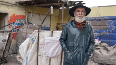 Tahsin Uzun, üzerinde deri ceketi ve kafasında fötr şapkası ile poz veriyor. Arkasında "çekçek" olarak adlandırılan tekerlekli çuvallar var. 