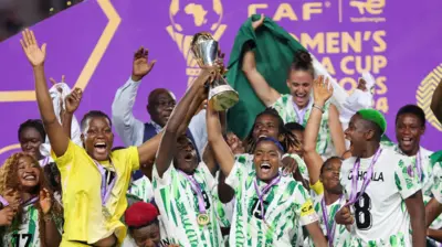 Un groupe de joueuses et d'officiels célèbre la victoire du Nigeria à la Coupe d'Afrique des Nations féminine. La capitaine Rasheedat Ajibade brandit le trophée, entourée de la gardienne Chiamaka Nnadozie et de l'attaquante Asisat Oshoala, tandis qu'Ashleigh Plumptre, drapée dans le drapeau nigérian, se tient à l'arrière-plan.