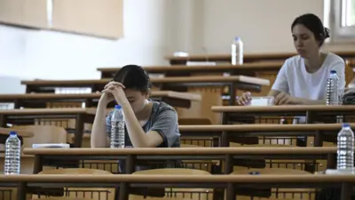 Sınav olan iki kızçocuğu aralıkla sıralarda oturuyor, yanlarında pet şişede sular görünüyor. 