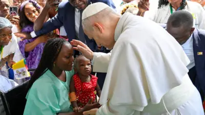 Pope Leo XIV wear white robe raise im hand dey pray for woman wey carry her pikin for leg. Oda pipo dey di background
