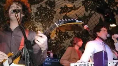 The Killers performing in a dark club in 2003. A man wearing a white shirt  sings into a microphone, while a guitarist with long curly hair sings along and a keyboardist plays in front of a wall decorated with a psychedelic design.