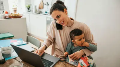 Bilgisayar başında bir kadın, kucağında bebek, kulağında telefon var
