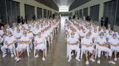 Presuntos miembros de pandillas, vestidos de blanco, con la cabeza rapada, mascarillas y atados con cadenas y sentados en sillas plásticas en el pasillo central de uno de los módulos del Centro de Confinamiento del Terrorismo (Cecot) en Tecoluca, El Salvador, asisten a una audiencia masiva en su contra el 20 de abril de 2026. (Reuters, foto proporcionada por una tercera parte)