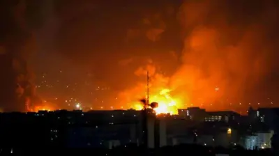 暗い建物群の向こうでオレンジ色の大きな火の手が複数上がり、煙が夜空に立ち上っている
