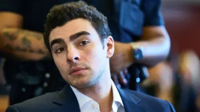 A head and shoulders shot of Luigi Mangione, in a dark jacket and white shirt. A uniformed officer with a tattoo on the arm stands behind him