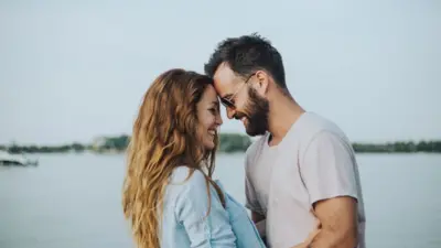 A man and a woman smile as they stand facing each other in front of a water body