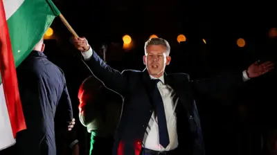 Magyar'ın sağ elinde macaristan bayrağı var, üzerinde koyu renk bir takım elbise ve kravat var.