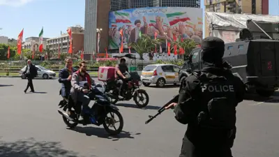 یکی از میدانهای تهران در حالیکه مامور پلیسی که صورت خود را پوشانده و اسلحه در دست دارد ایستاده. عابران و دو موتور سوار در حال گذار از خیابان هستند.