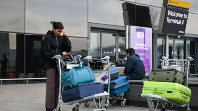 Woman and man wait with brightly coloured suitcases on trolleys outside Departures at Heathrow Terminal 4