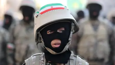 Um jovem usando balaclava, uniforme camuflado e capacete com a bandeira do Irã olha para fora do enquadramento, com figuras militares desfocadas ao fundo