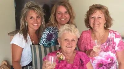 Anita LeBrun, de 82 anos, veste uma blusa com estampa floral rosa-choque e um corsage; ela sorri enquanto ergue uma taça de vinho branco, cercada pelas mulheres de sua família.