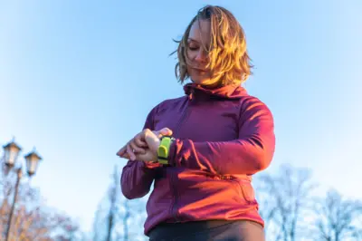 Kobieta w czerwonym polarze patrzy na zielony smartwatch. Ma lekko zmartwiony wyraz twarzy. Za nią widać korony drzew i latarnię. 