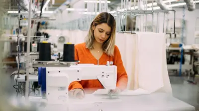 Dikiş makinesinde çalışan turuncu kıyafetli bir kadın. Arkada bir fabrika ortamı var.