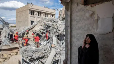 27 मार्च को तेहरान में हवाई हमले में क्षतिग्रस्त हुई एक रिहायशी इमारत के मलबा हटाते इमरजेंसी वर्कर्स और पास में फोन पर बात करती एक महिला.