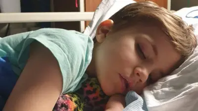 Roo, uma criança pequena deitada em uma cama de hospital, segurando um brinquedo