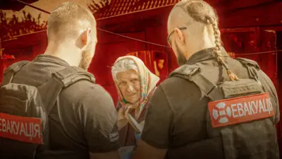 Двоє чоловіків у бронежилетах із написом «ЕВАКУАЦІЯ» спілкуються з літньою жінкою, яка тримає документи та стривожено дивиться повз камеру. Сцена виглядає як допомога цивільним у зоні небезпеки. 