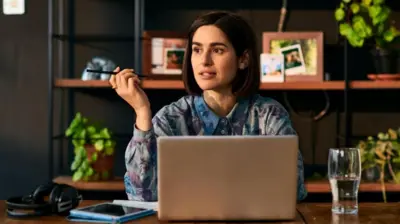 Bir kadın düşünceli bir şekilde laptop başında oturuyor, ofise benzer bir alanda masasının üzerinde bardakta su, kulaklıklar ve tableti var. Arka planda fotoğraf ve çiçeklerin olduğu bir raf.