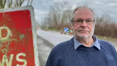 Phil Barlow standing next to a red sign warning motorists to stop at the traffic lights. He is wearing a knitted navy jumper with a navy shirt underneath, and has grey hair and glasses on. Cones are pictured behind Barlow.