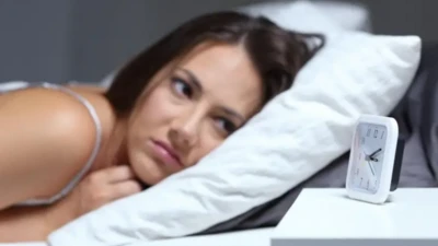 Leżąca w łóżku kobieta patrzy zmartwionym wzrokiem na budzik stojący na białej szafce. 