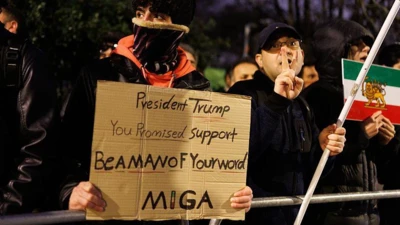 Несколько людей стоят за заграждением, один человек держит флаг Ирана, молодой человек в центре кадра держит плакат с надписью President Trump, You promised support, be a man of your word MIGA («Президент Трамп, вы обещали поддержку, будьте человеком слова MIGA». Последняя аббревиатура, по всей видимости, означает Make Iran great again, т.е. «сделаем Иран снова великим»).