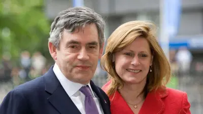Gordon and Sarah Brown. He has grey hair and is looking at the camera. he is wearing a blue suit with a white shirt and purple tie. Sarah is looking off camera and is wearing a red jacket.