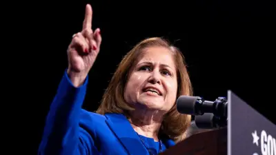 Hindistonlik 61 yoshli G’azala Hoshimiy ko‘k kostyumda, yelkasigacha bo‘lgan qora sochli, minbarda mikrofonga gapirmoqda. Uning o‘ng qo‘li yuqoriga ko‘tarilib, ishora barmog’i yuqoriga ko‘rsatilgan.