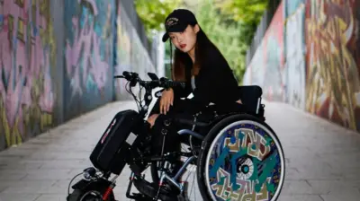 양쪽 벽에 그래피티가 그려진 콘크리트 길 위에 검은 모자를 쓴 긴 검은 머리의 여성이 휠체어에 앉아 있다. 휠체어 바퀴에는 그래피티가 그려져 있다. 