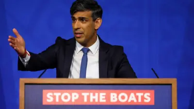 Sunak gesticulando em discurso; na frente, no palanque, aparece frase: Stop the boats (Parem com os barcos)