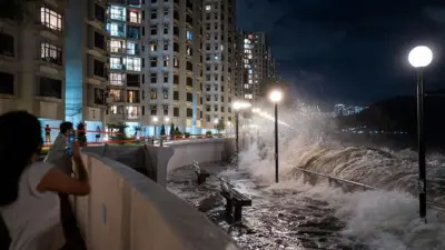 夜間一個靠海的住宅區，畫面右側有巨浪拍打岸邊護欄，海水湧入步道，把長椅、欄杆和燈柱幾乎完全淹沒。有一名女子正拿著手機拍照。背景是一排高層住宅大廈。