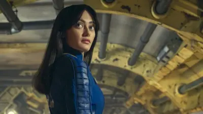 A young woman with long black hair stands in an artificially lit, cylindrical corridor with various pipes supported by bright yellow struts along its length She's looking back over her shoulder with an impassive expression. She wears a close-fitting, dark blue leather jumpsuit with panelling down the arms.