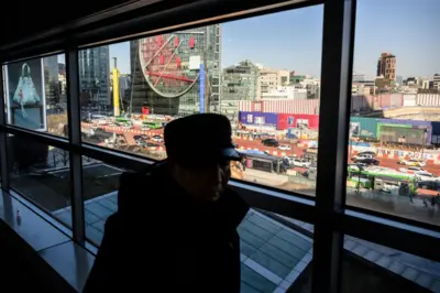 유리창 너머로 서울 도심의 번화한 거리와 고층 건물이 보인다. 창문 안쪽에는 모자를 쓴 한 남성이 서 있고, 바깥에는 차량과 버스가 도로를 따라 이동하고 있다.