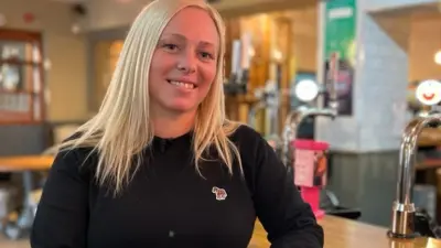 A young woman with long, blonde hair wearing a black pullover smiles as she sits at the bar of the pub she manages. The lights of the pumps on the bar twinkle behind her. 