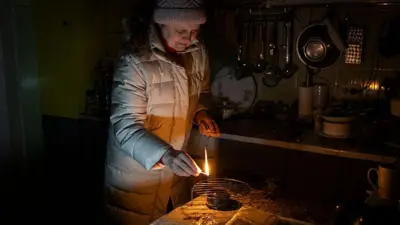 Женщина зажигает "окопную" свечу в темной кухне. 