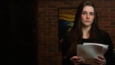 Michele olhando sério para cama, em sala de casa, segurando papelada