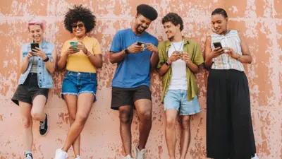 Em fotografia de banco de imagens, cinco jovens de diferentes perfis aparecem apoiados contra uma parede desgastada enquanto usam smartphones e interagem entre si. Eles usam roupas coloridas, sorriem e observam as telas dos celulares.