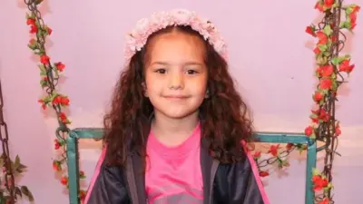 Hind Rajab sorri para a câmera. Ela tem cabelos longos, castanhos e ondulados, e usa uma coroa de flores na cabeça. Também veste uma camiseta rosa e uma jaqueta preta com zíper. Ela está sentada em uma cadeira decorada com guirlandas de flores vermelhas.
