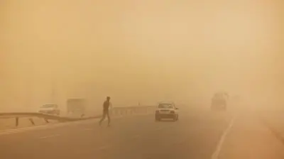 Ọkùnrin kan fẹ́ sọdá ojú pópó, àwọn ọkọ̀ ń kọjá, kùrukùru bo ojú ọjọ́