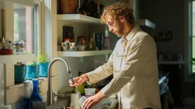 Homem com cabelo ondulado, vestindo uma camisa bege, lavando pratos