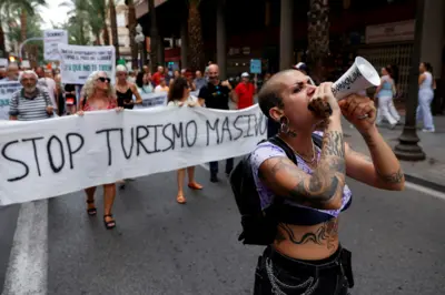 A demonstrator with tattoos uses a megaphone as people protest against mass tourism on a street in Alicante, Spain, July 13, 2024. The banner reads "Stop turismo masivo" ("Stop mass tourism")