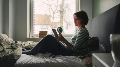 Kobieta w średnim wieku siedzi na łóżku. W dłoniach trzyma telefon komórkowy i przenośny wentylator. Przez okno za nią widać czerwone dachy domów i drzewa. 