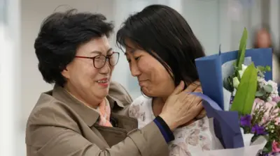Two women, one older and one younger, embrace. They look like they are crying with Joy, and one is also holding a bunch of flowers