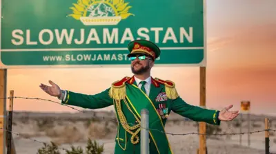 Un hombre vestido en un traje militar verde de gala con decoraciones, gafas de sol y gorro militar está parado frente a una valla que lee: "República de Solwjamastan". En el fondo se divisa un desierto y un cielo con los colores del atardecer
