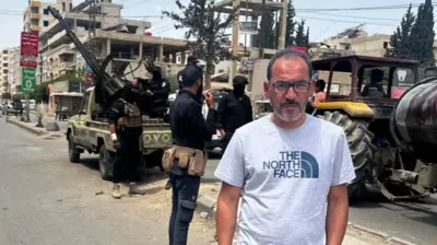 Feras Kilani stands on a street in Damascus, Syria. In the background, Syrian security forces are deployed with a truck-mounted anti-aircraft gun (7 May 2025) 