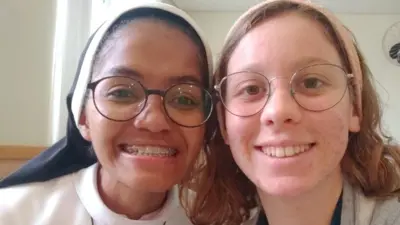Fotografia colorida mostra duas mulheres de óculos sorrindo para a foto. A da esquerda usa um hábito de freira e a da direita usa uma passadeira rosa no cabelo
